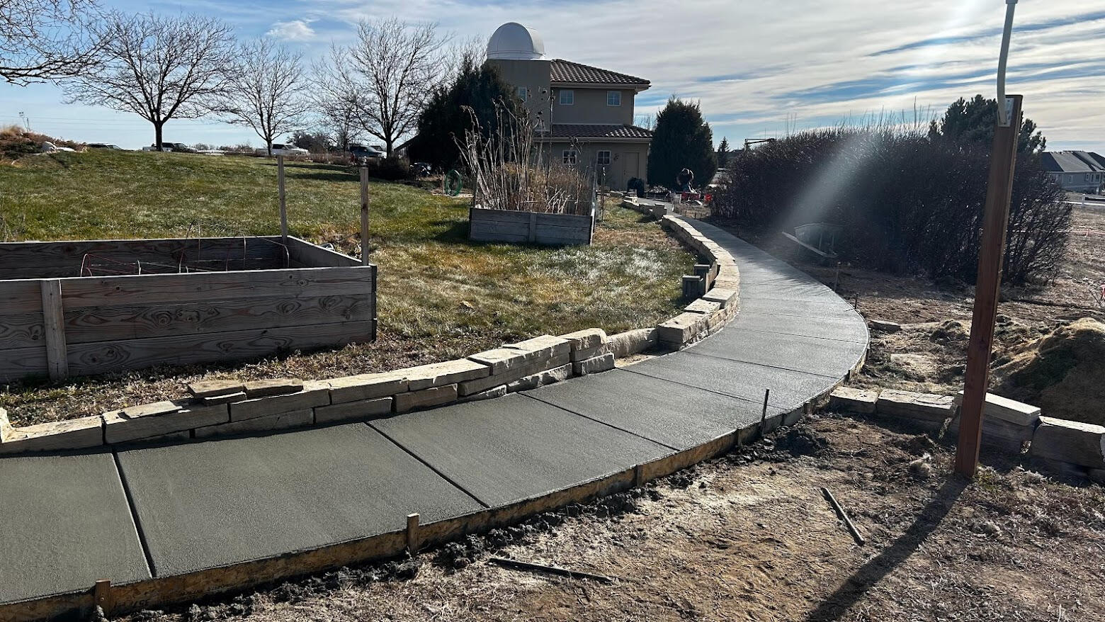 Curved concrete sidewalk in Severance, CO with broom finish, bordered by stone retaining wall in residential yard.