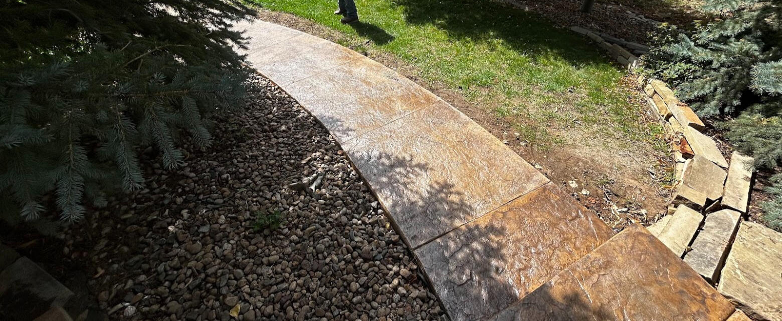 Stamped concrete walkway with natural stone texture curving through landscaped yard, bordered by river rock and trees