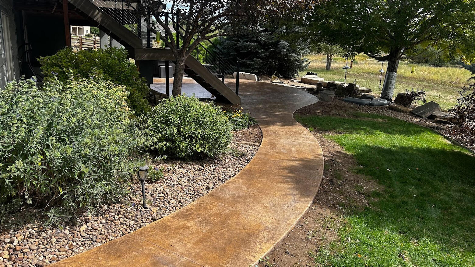 Stamped concrete walkway with natural stone texture winding through landscaped yard with trees, plants, and rock borders.