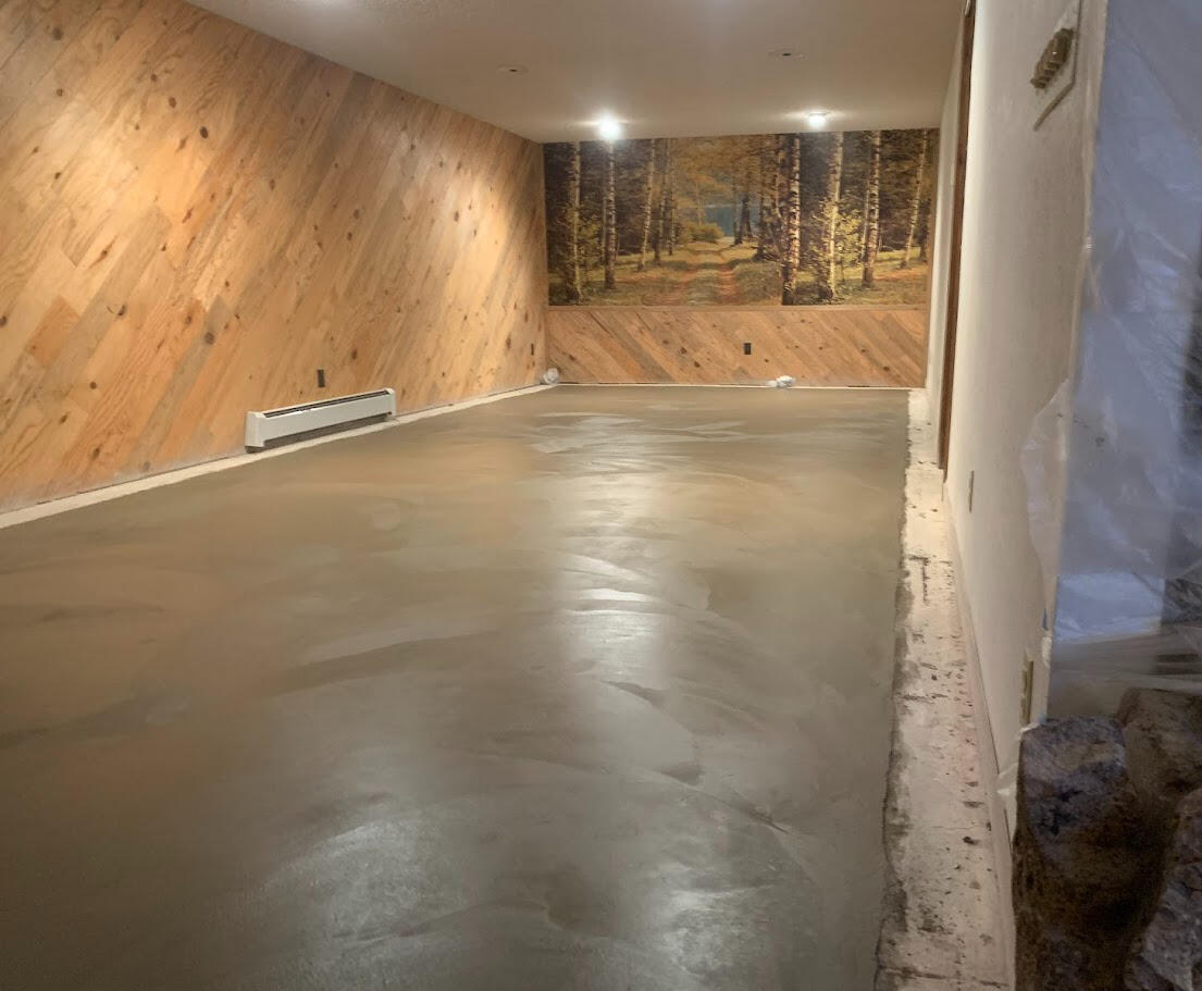 Freshly finished concrete floor in narrow basement room with wood-paneled wall and forest mural accent in Wellington, CO